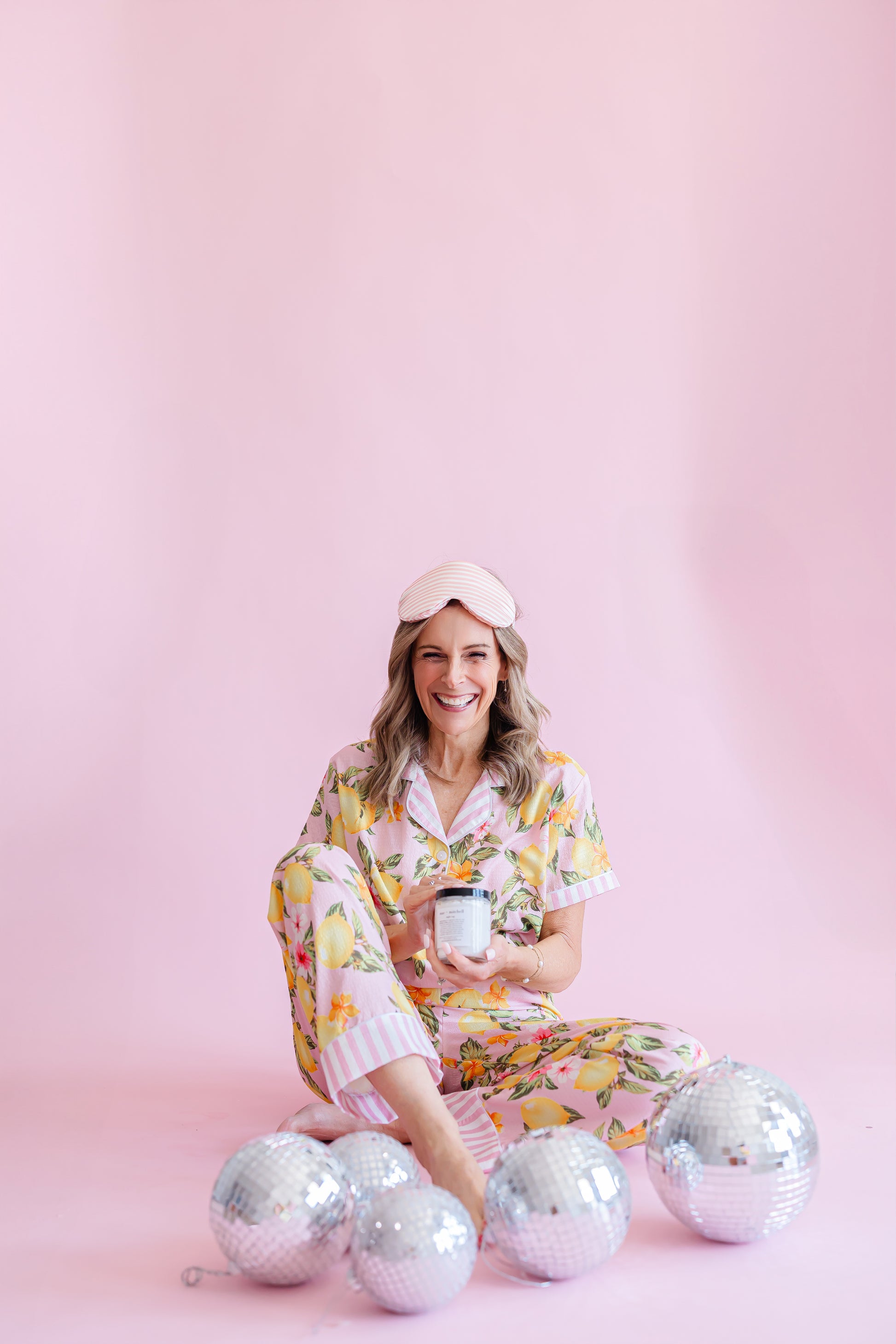woman in pajamas wearing sleep mask holding mac + mitchell nightcap magnesium cream, pink background